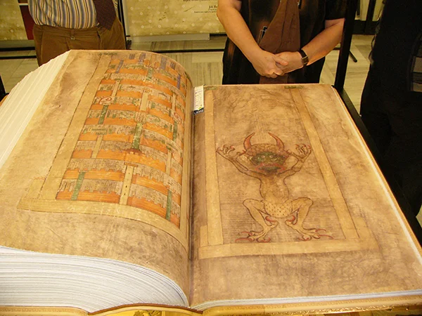 広げられた中世の巨大な古書(コデックス・ギガス)の右ページに描かれた、角と鋭い爪を持つ悪魔の細密な彩色挿絵。