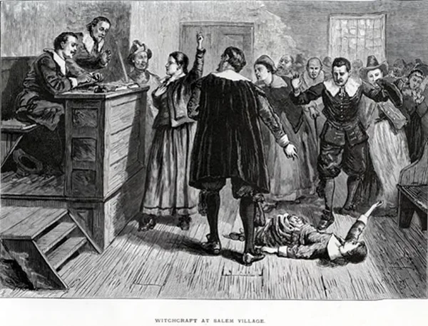 17世紀アメリカのセーラム村における魔女裁判の場面を描いた白黒版画。床に倒れた女性を周囲の人々が指差して非難しており、下部に「WITCHCRAFT AT SALEM VILLAGE」と記されている。